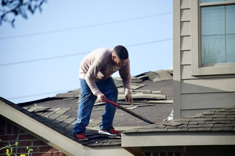 A worker installing light-colored metal roofing panels on a residential home under a blue summer sky.